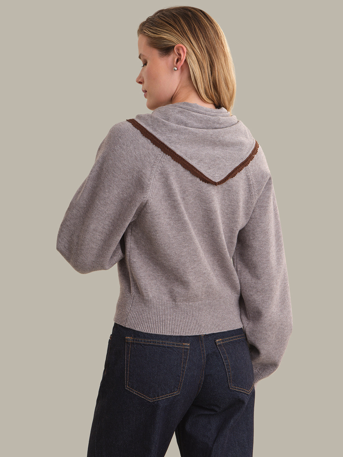 A woman shown from the back wearing a light gray knit cardigan with a brown textured hood trim, paired with dark blue jeans, against a solid light gray background.