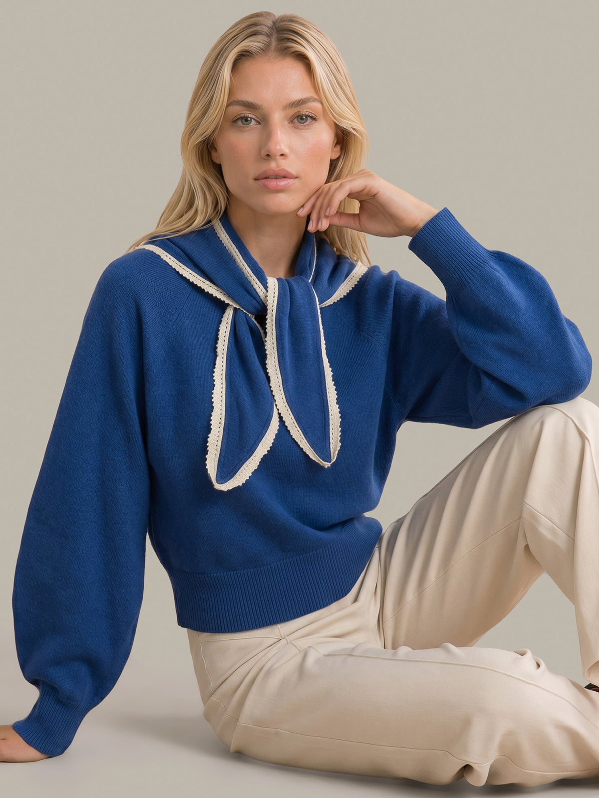 A woman wearing a royal blue knit sweater with a large cream bow detail at the neckline, paired with beige pants, posing against a solid light gray background.