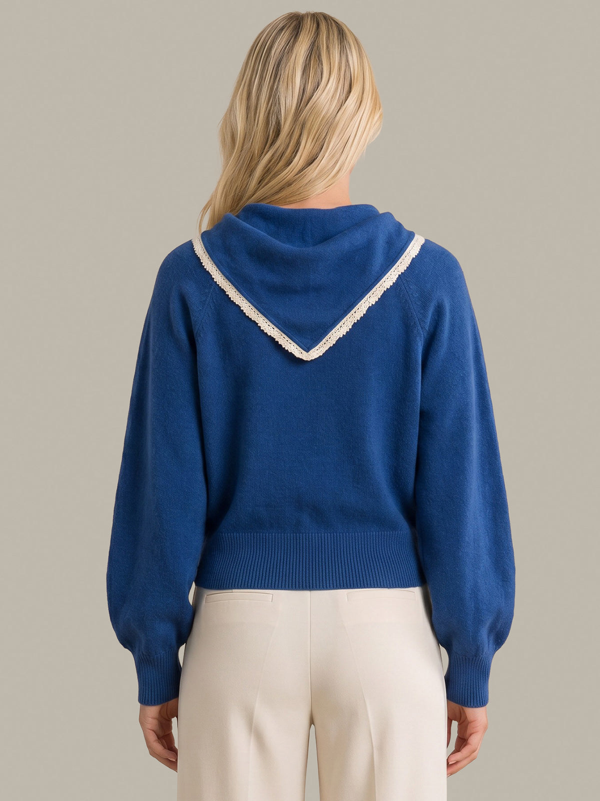 A woman shown from the back wearing a royal blue knit sweater with a white lace-trimmed V-shaped neckline detail, paired with cream-colored pants, against a solid light gray background.