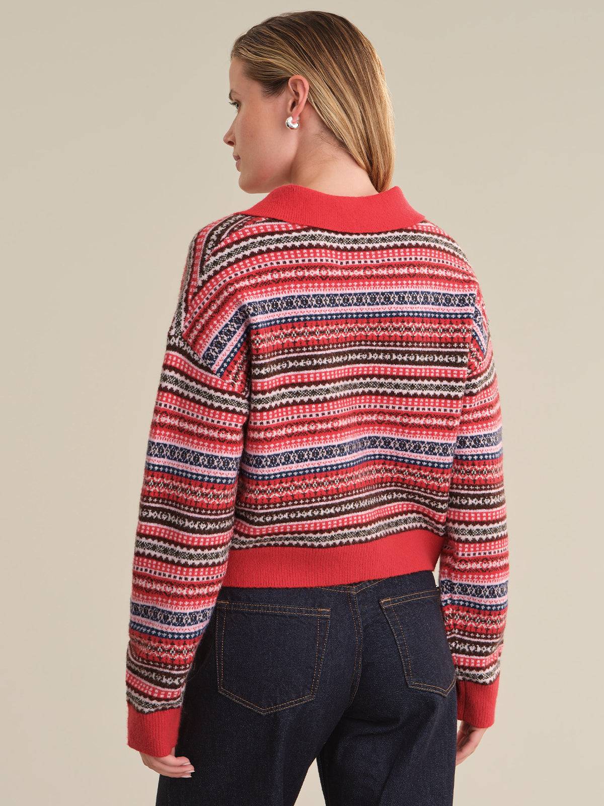 A woman shown from the back wearing a red, navy, and cream striped knit sweater with dark blue jeans, set against a solid beige studio background.