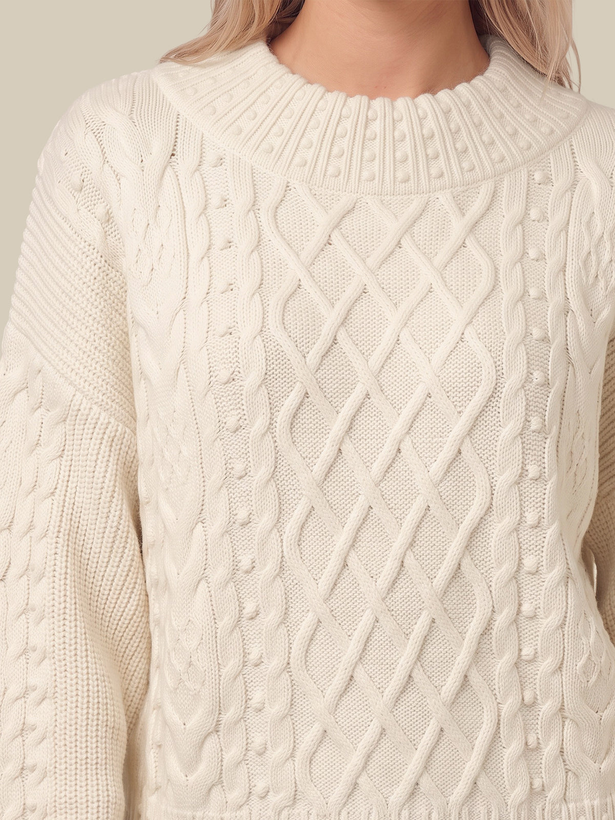 A close-up of a cream-colored cable-knit sweater with a textured diamond pattern on the front, worn by a model.