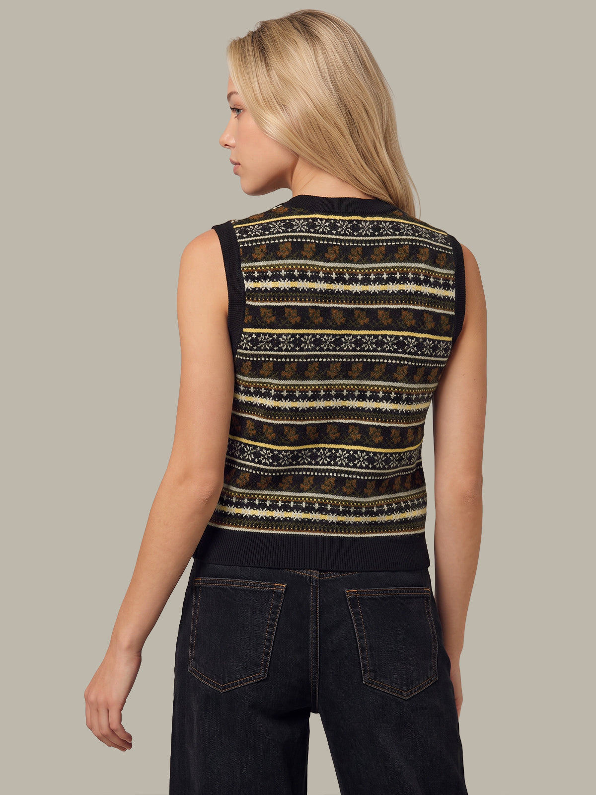A woman shown from the back wearing a sleeveless knit top with a black base and horizontal geometric patterns in beige and brown, paired with dark jeans against a solid light gray background.