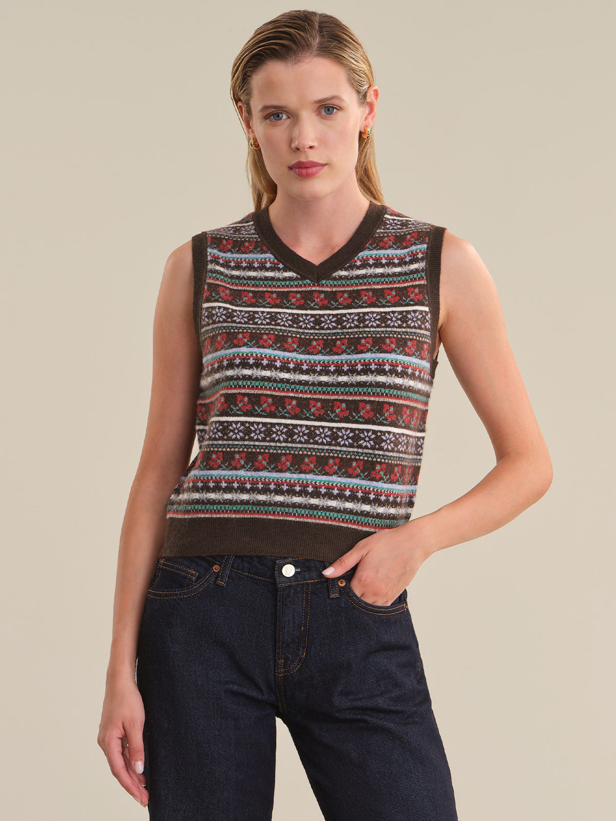 A woman modeling a sleeveless knit sweater vest with a brown, red, and white Fair Isle pattern, paired with dark blue jeans, against a solid beige background.
