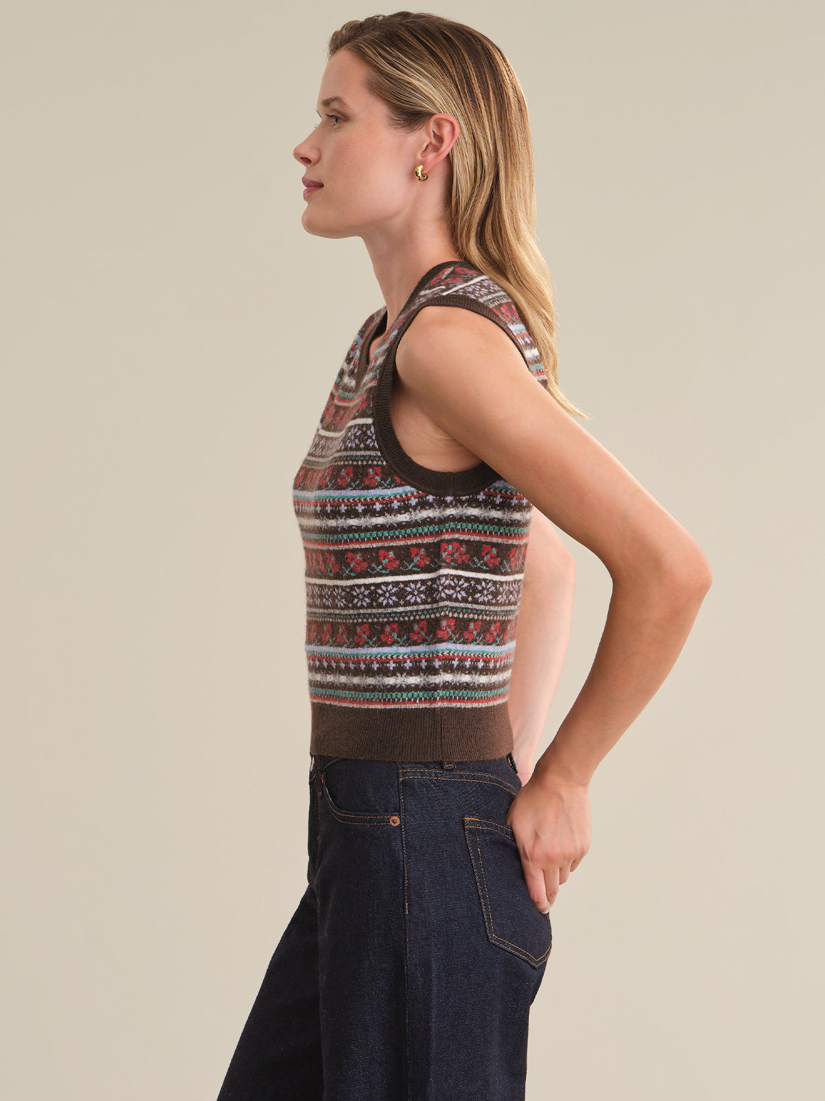 A woman shown in side profile wearing a sleeveless knit top with a multicolor Fair Isle pattern and dark blue jeans, against a solid beige studio background.