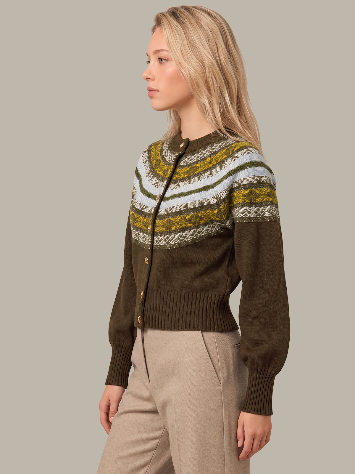 A woman wearing a dark olive green knit cardigan with a colorful patterned yoke in white, olive green, and mustard yellow, paired with beige trousers, set against a solid light gray background.