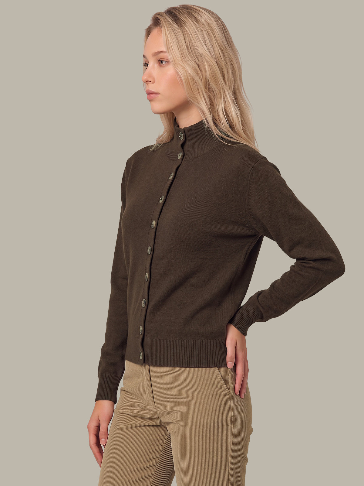 A woman modeling a dark brown button-up knit cardigan sweater with a mock neck, paired with beige pants, against a solid light gray background.