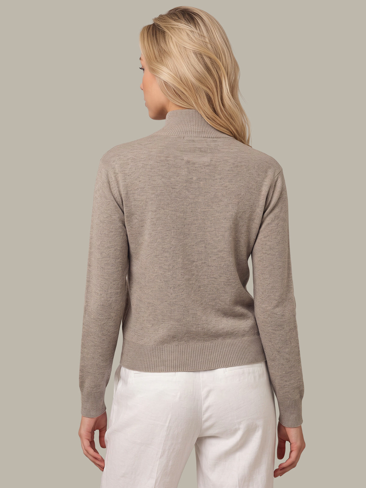 A woman shown from the back wearing a taupe-beige ribbed mock neck sweater and white pants, set against a solid light gray studio background.