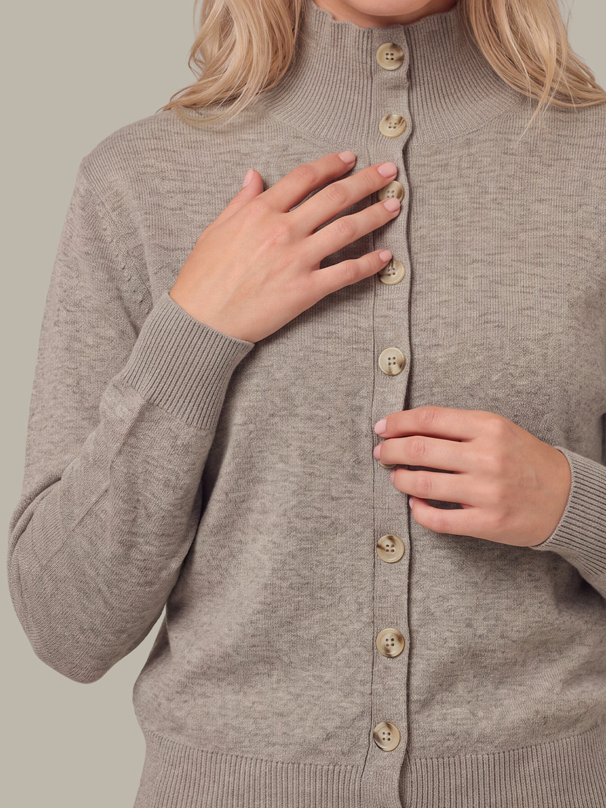A close-up of a woman wearing a taupe knit cardigan sweater with a mock neck and beige buttons, with her hands resting on the front of the sweater.