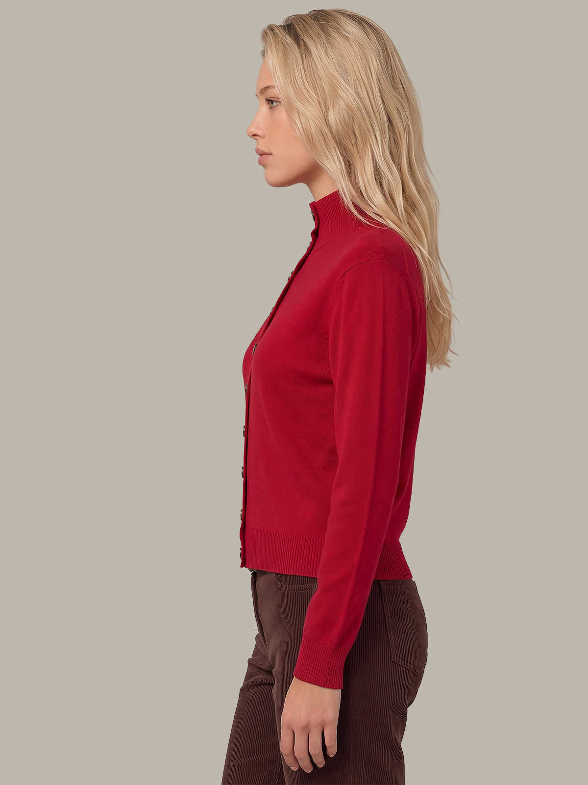 A woman in profile wearing a red mock-neck button-up cardigan and brown pants, photographed against a solid light gray studio background.