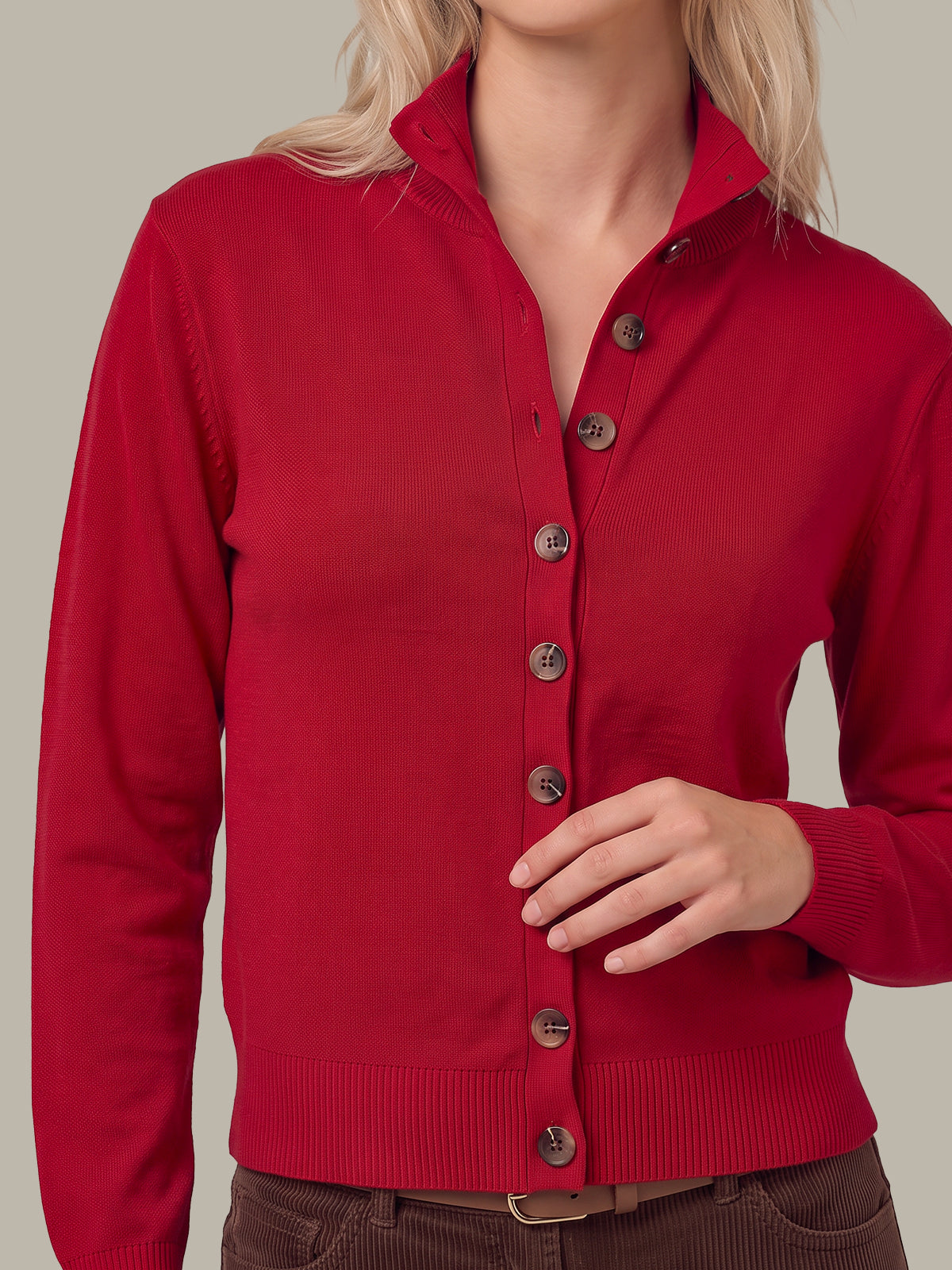 A woman wearing a bright red knit cardigan sweater with a stand collar and dark buttons, paired with brown corduroy pants, against a solid light gray background.