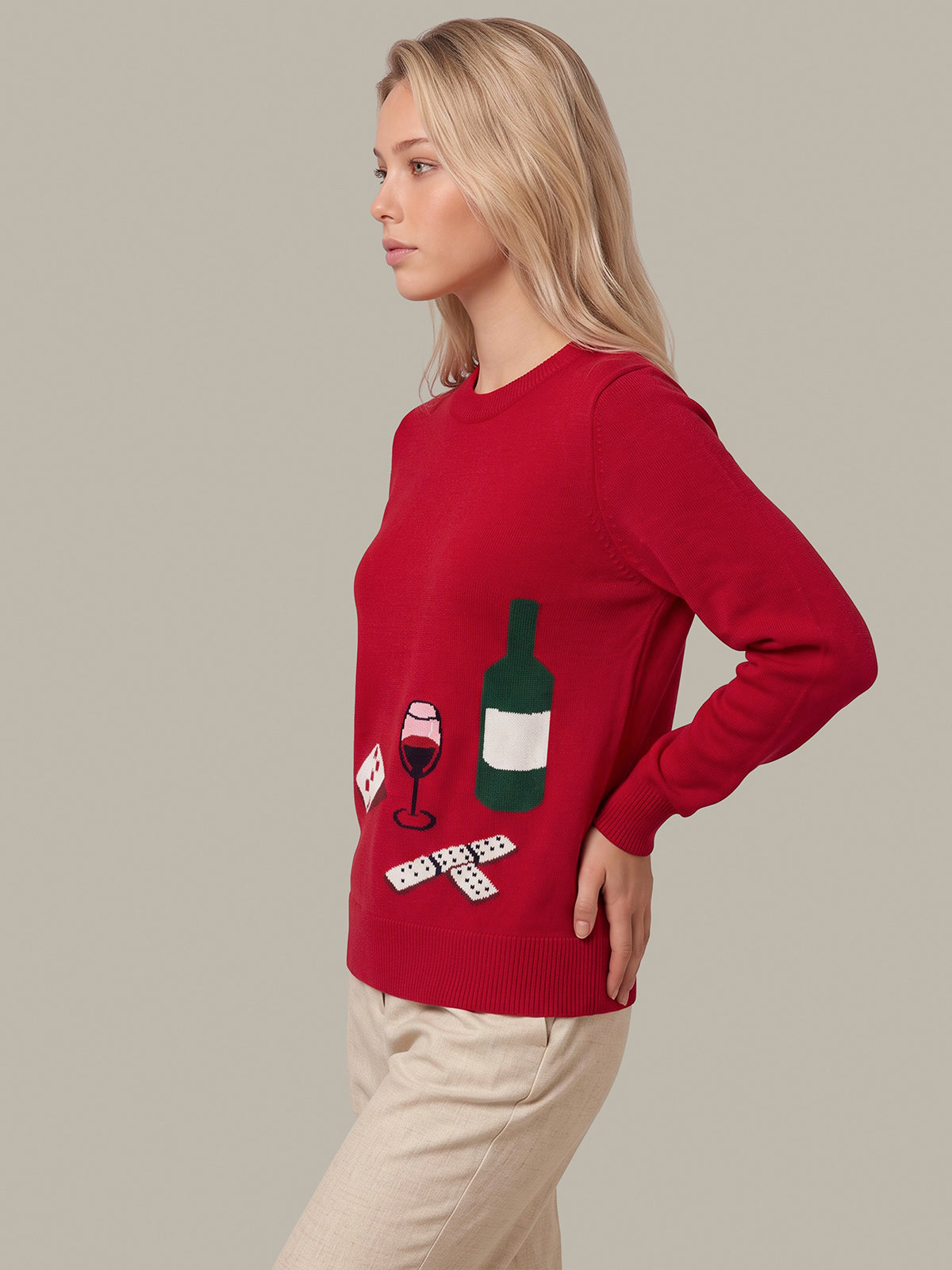 A woman wearing a red crewneck sweater with a wine-themed graphic print, paired with beige trousers, photographed against a solid light gray studio background.