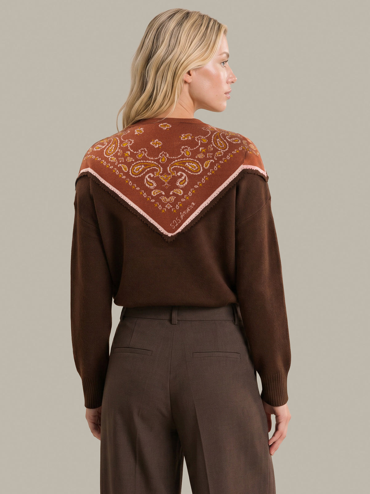 A woman shown from the back wearing a brown knit sweater with a large rust-orange paisley bandana yoke and dark brown trousers, set against a solid light gray background.