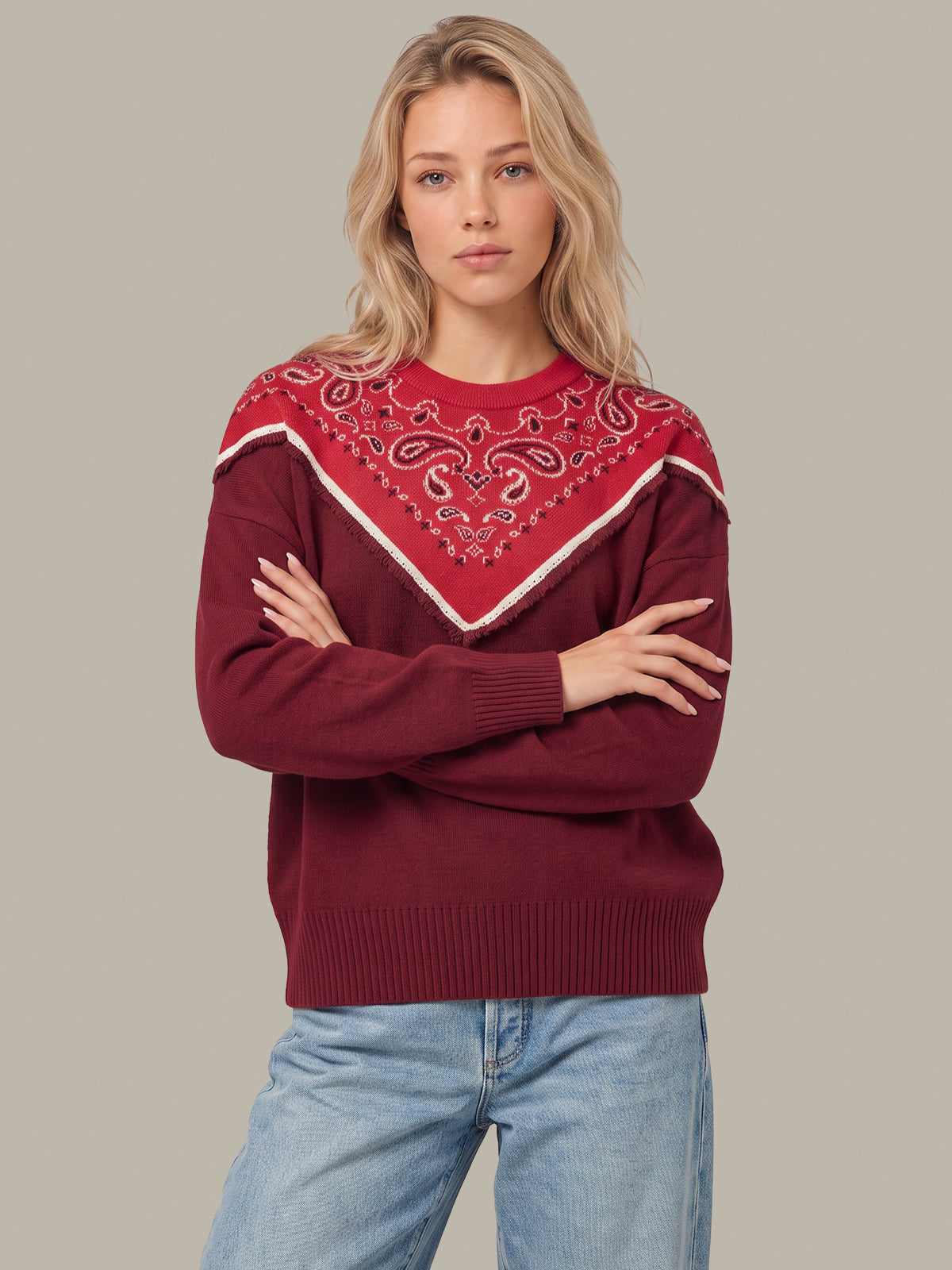 A woman modeling a burgundy knit sweater with a red and white paisley bandana pattern across the chest, paired with light blue jeans, against a solid gray background.