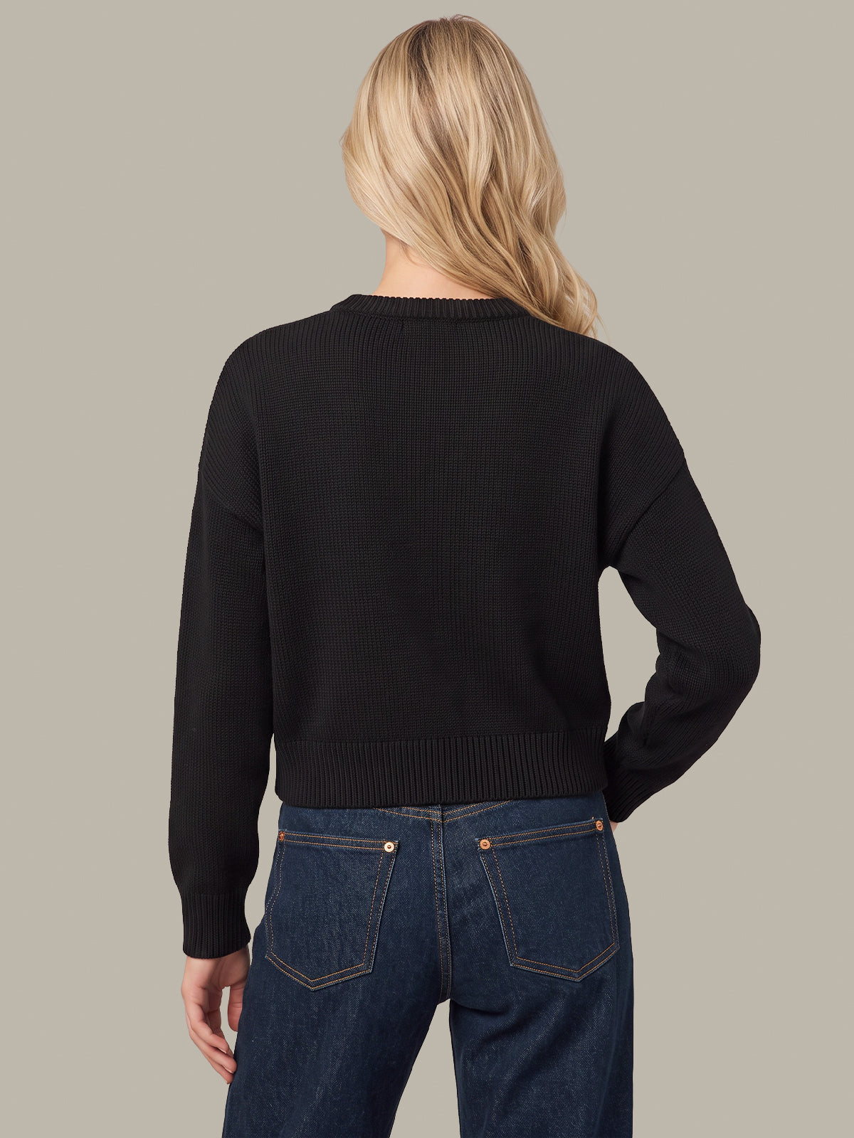 A woman viewed from the back wearing a black ribbed long-sleeve cropped sweater and dark blue jeans against a solid taupe background.