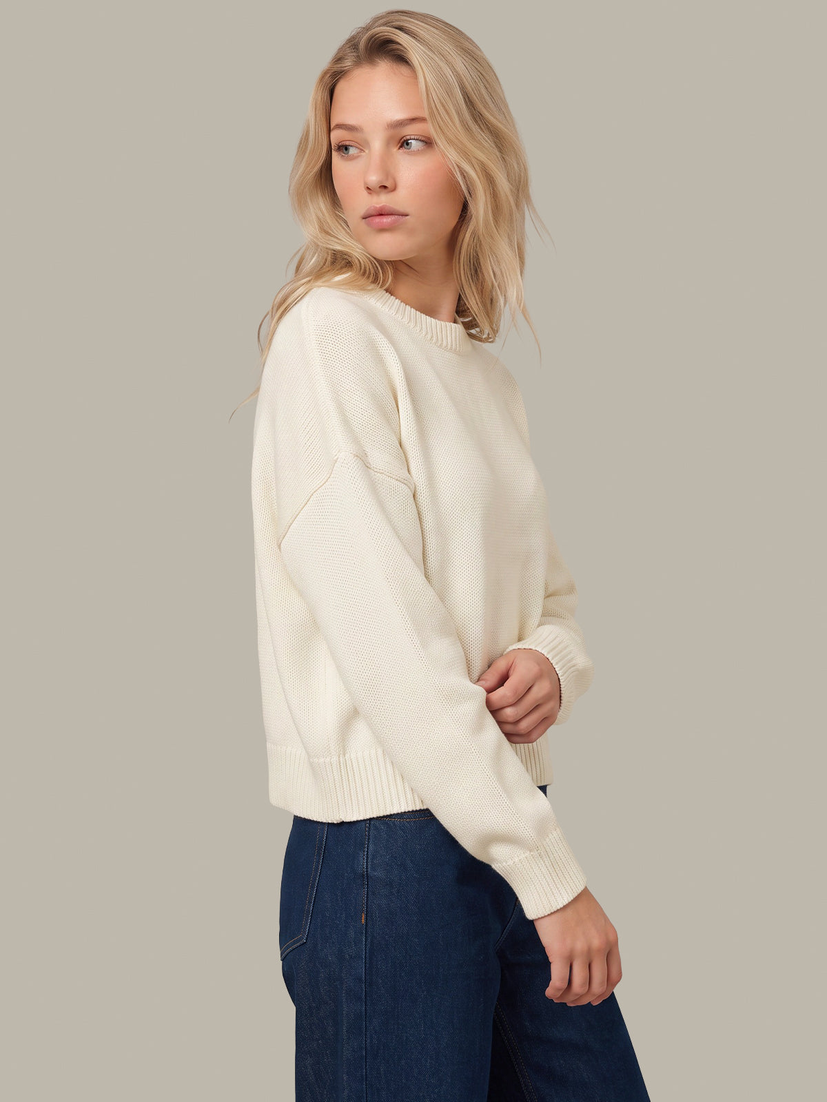 A woman wearing a cream-colored knit sweater and dark blue jeans, photographed from the side against a solid light gray background.
