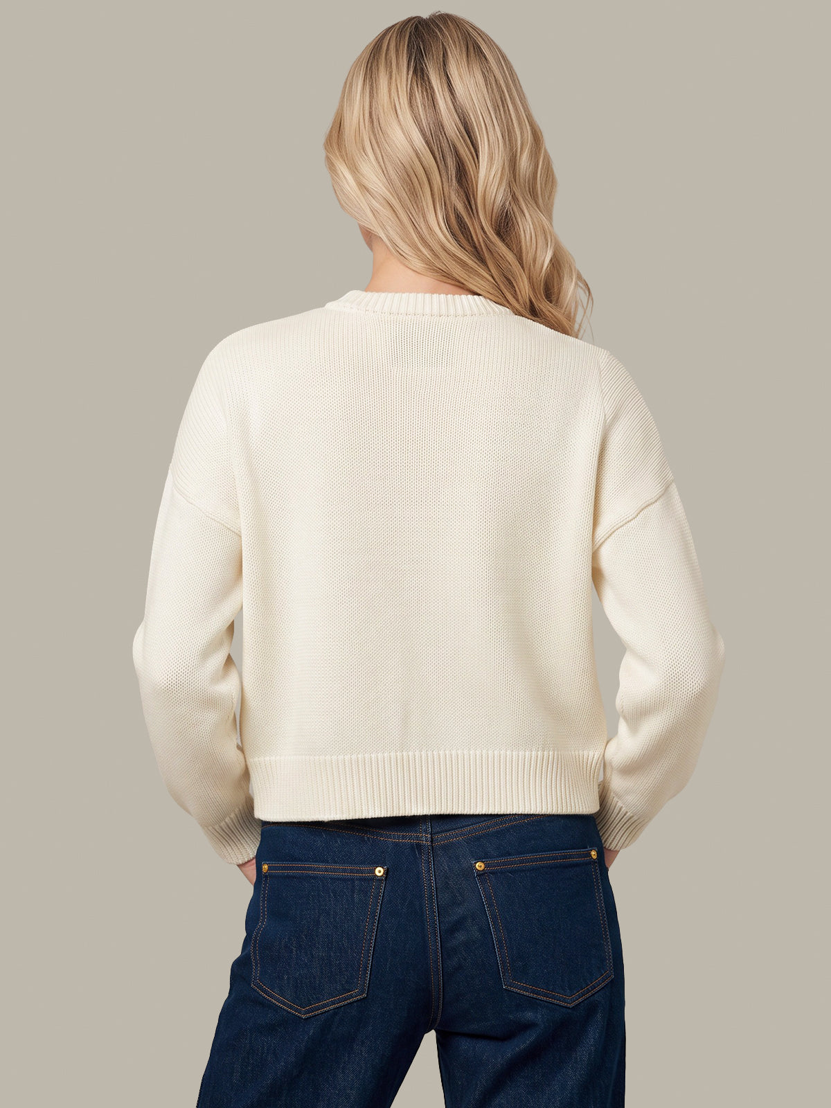 A woman viewed from behind wearing a cream-colored knit sweater and dark blue jeans, set against a solid light gray studio background.