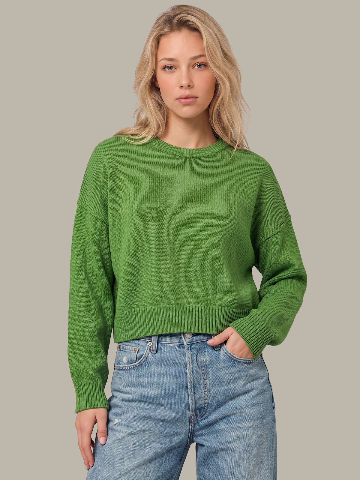 A woman wearing a bright green ribbed knit crewneck sweater and light wash blue jeans, standing against a solid neutral gray background.