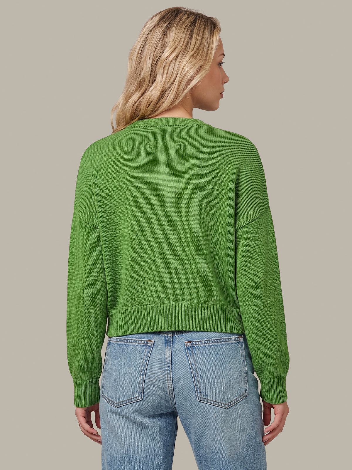 A woman shown from the back wearing a bright green cropped knit sweater and light blue jeans against a solid taupe background.