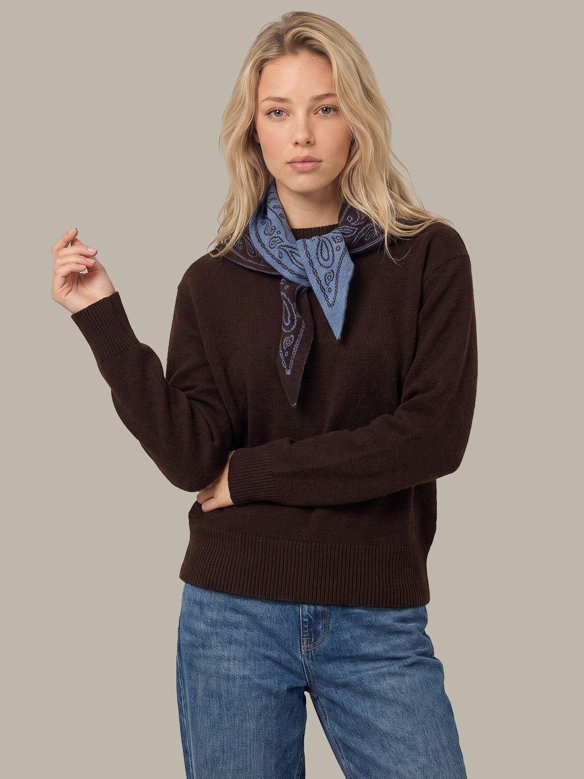 A blonde woman modeling a dark brown ribbed knit sweater, styled with a blue paisley bandana tied around her neck and blue jeans, against a solid light gray background.