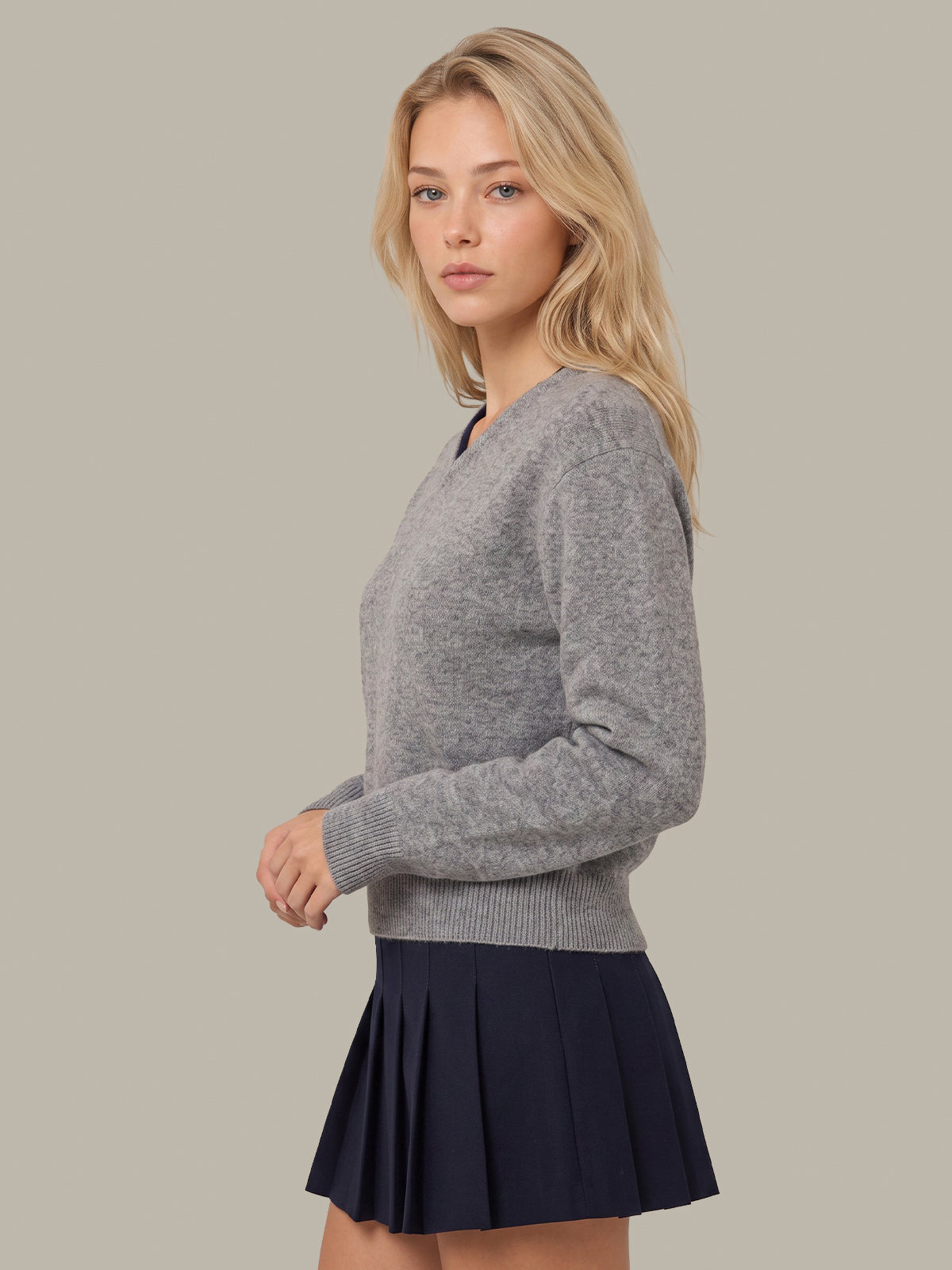 A blonde woman modeling a light gray v-neck knit sweater paired with a dark navy pleated mini skirt, set against a solid light gray studio background.
