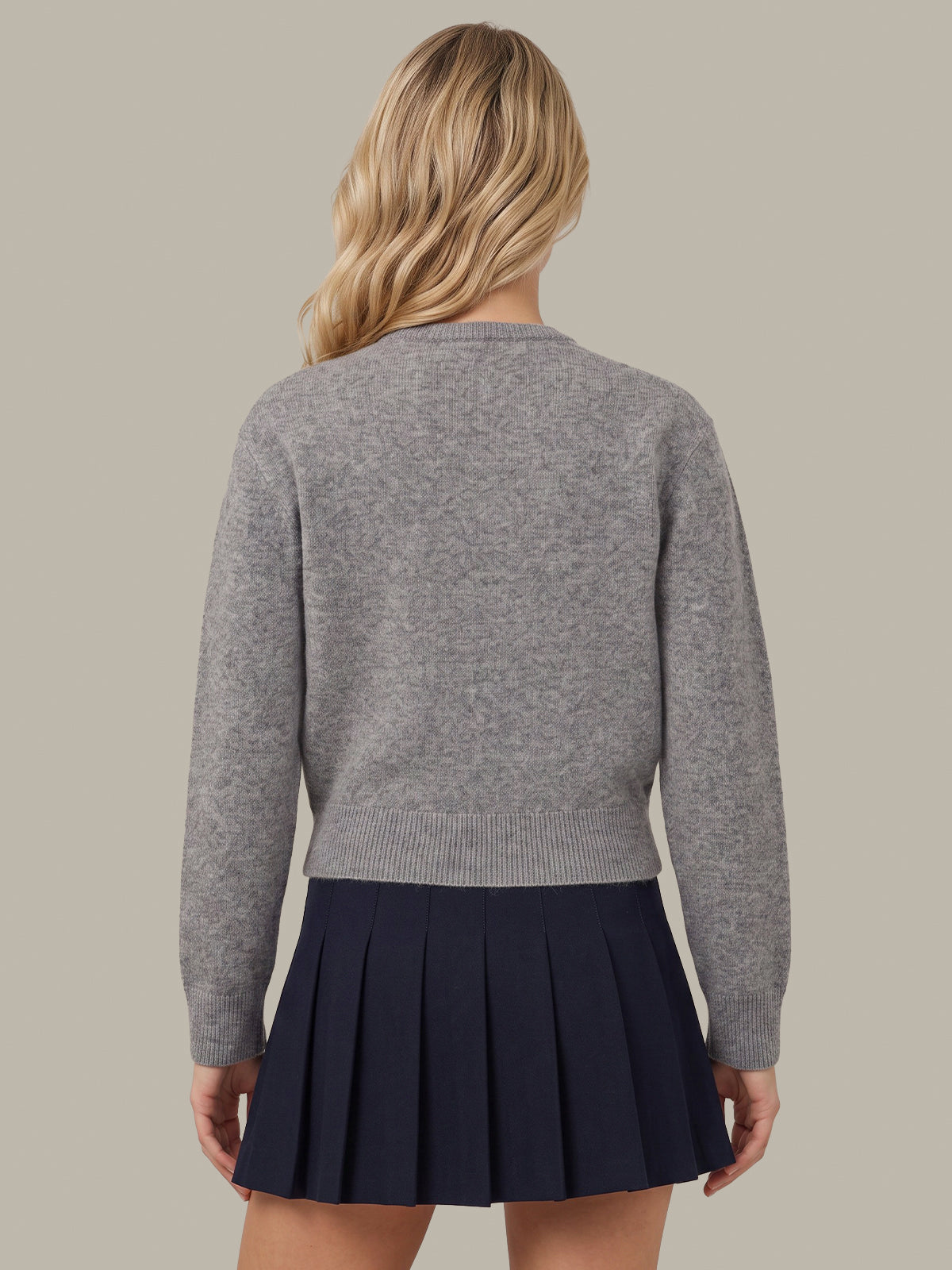 A woman shown from the back wearing a heather gray long-sleeve knit sweater and a dark navy pleated mini skirt, set against a solid warm gray studio background.