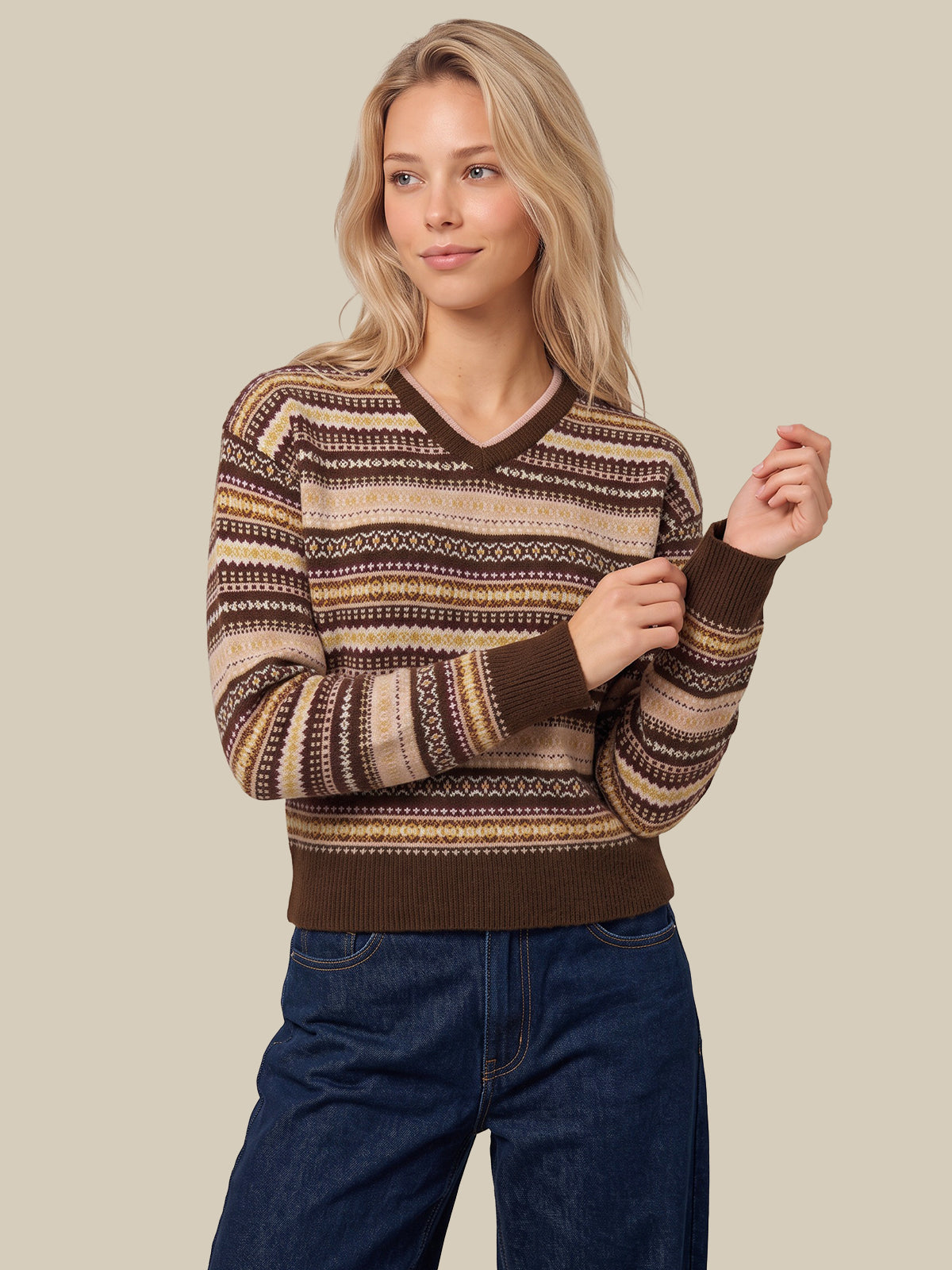 A woman wearing a brown and beige patterned knit sweater with a V-neck, paired with dark blue jeans, against a solid beige background.