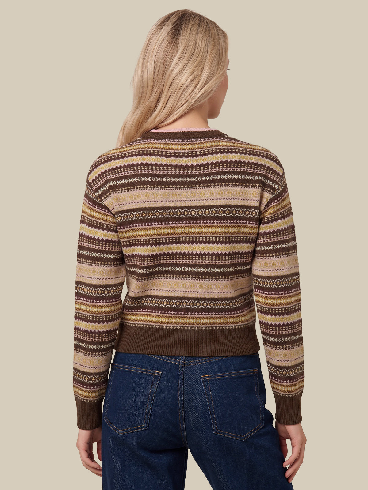 A woman viewed from the back wearing a brown, beige, and cream patterned knit sweater and blue jeans, set against a solid beige background.