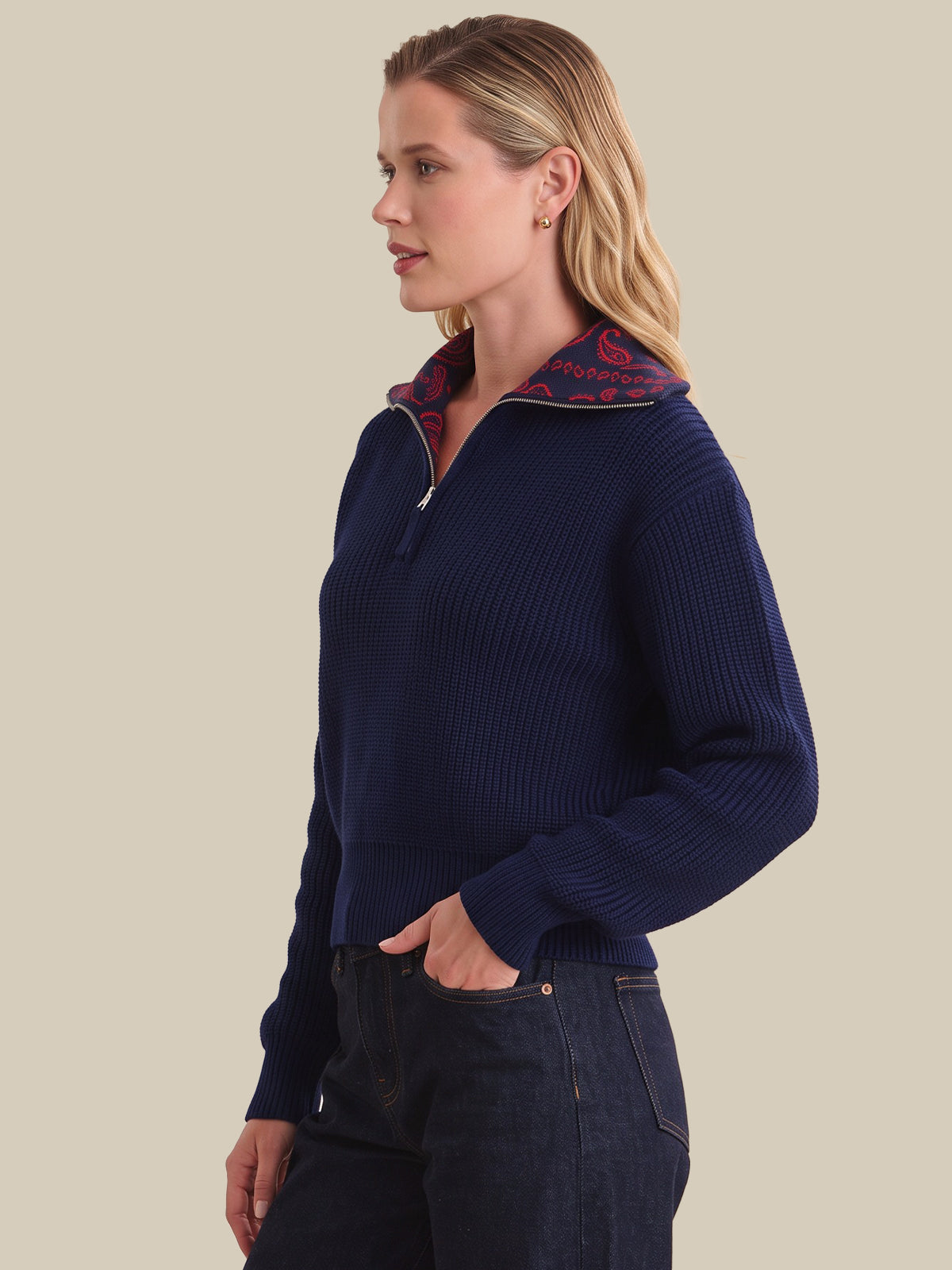 A woman shown in profile wearing a navy ribbed quarter-zip sweater with a patterned inner collar, paired with dark jeans, against a solid beige background.