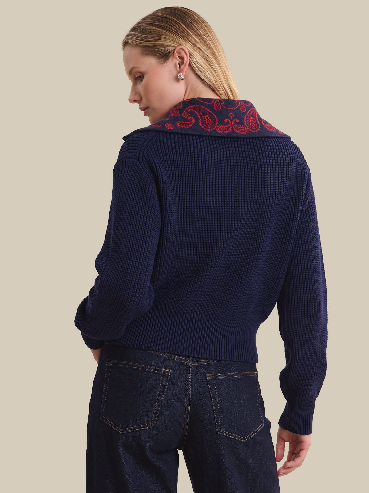 A woman shown from the back wearing a navy blue ribbed knit cardigan with a red paisley patterned collar, paired with dark blue jeans, against a solid beige background.