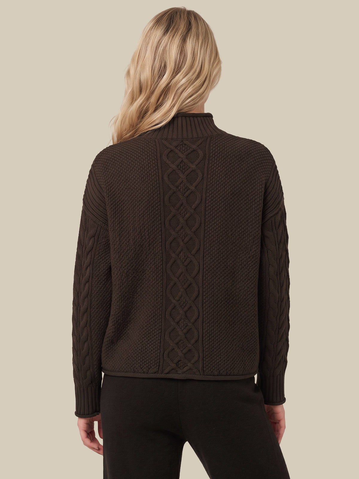 A woman viewed from the back wearing a dark brown cable-knit mock-neck sweater and black pants, set against a solid beige background.