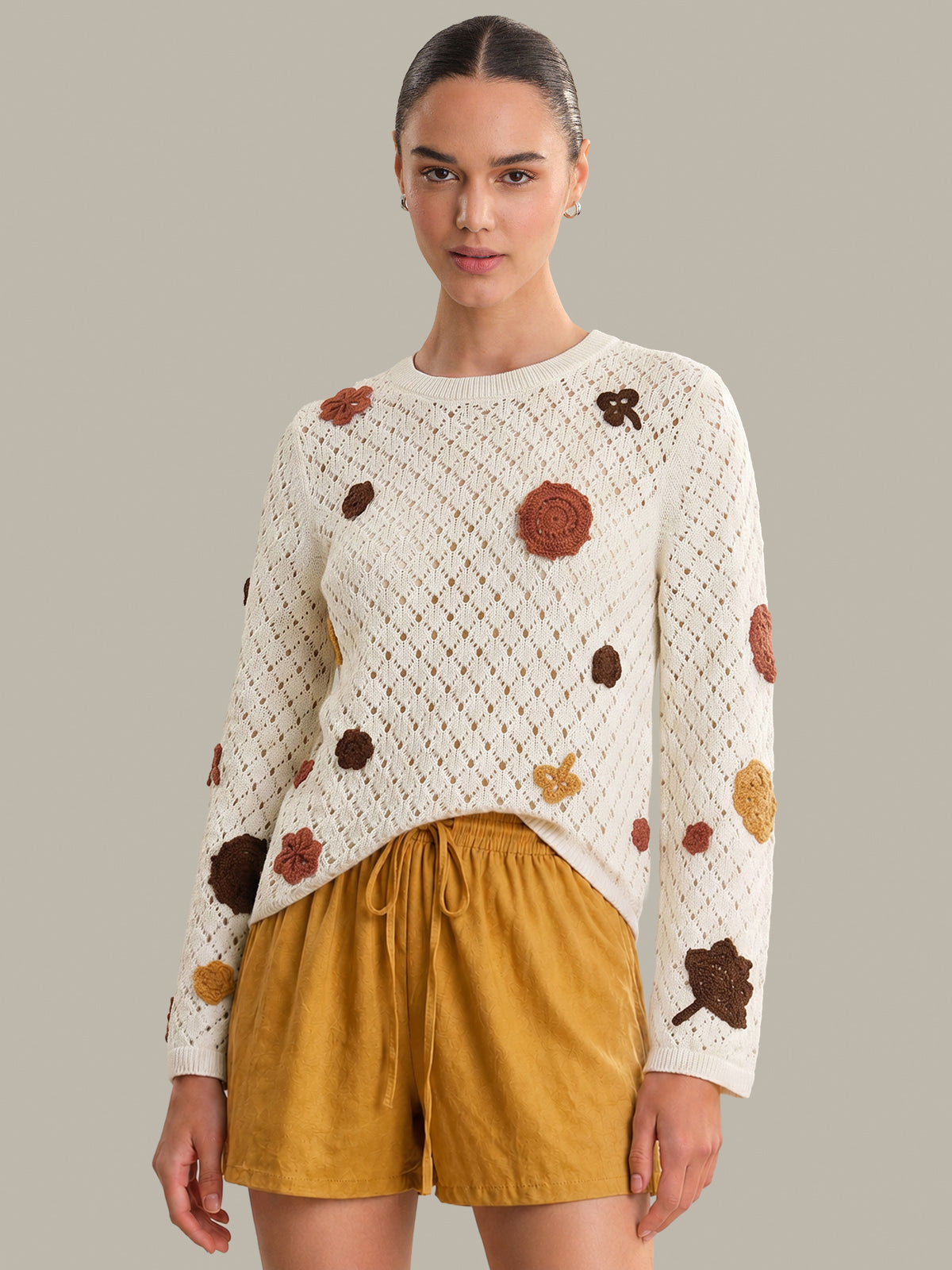 Woman wearing a white sweater with brown and gold floral patterns and mustard yellow shorts on a gray background