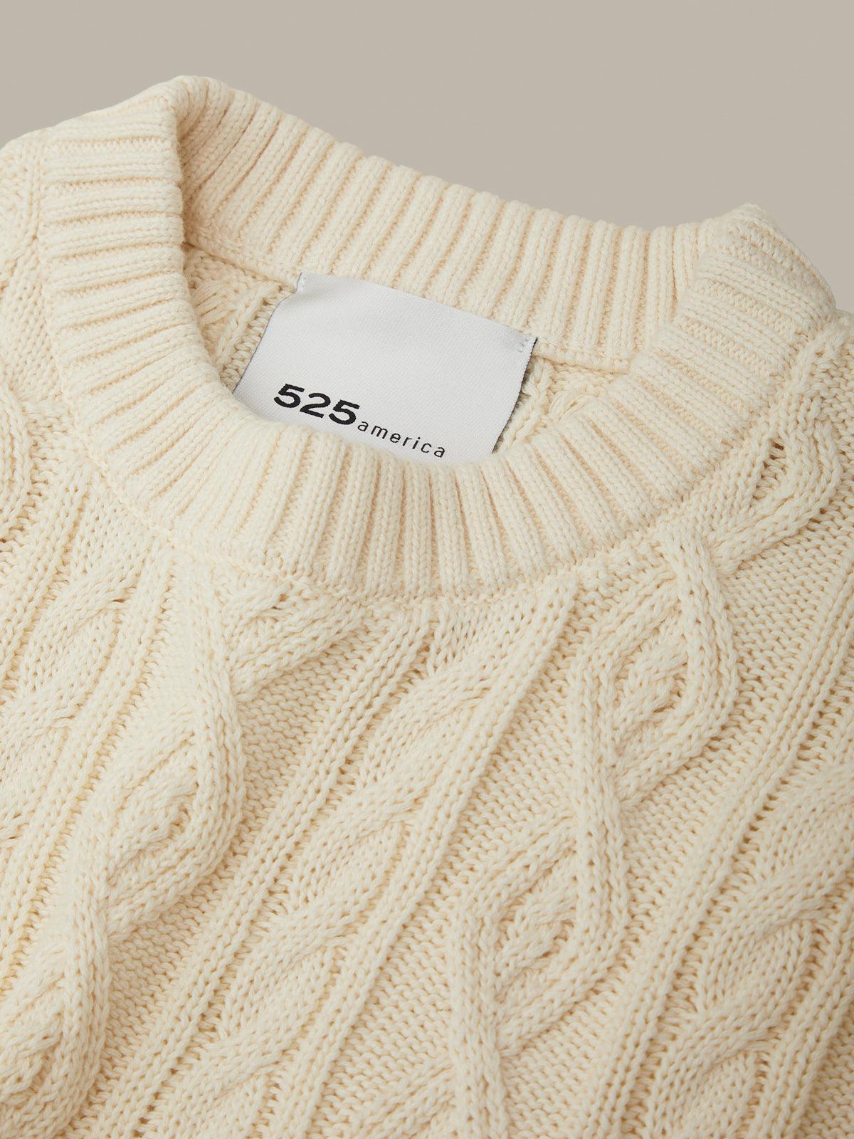 Close-up of a beige cable knit sweater with a brand tag on a neutral background