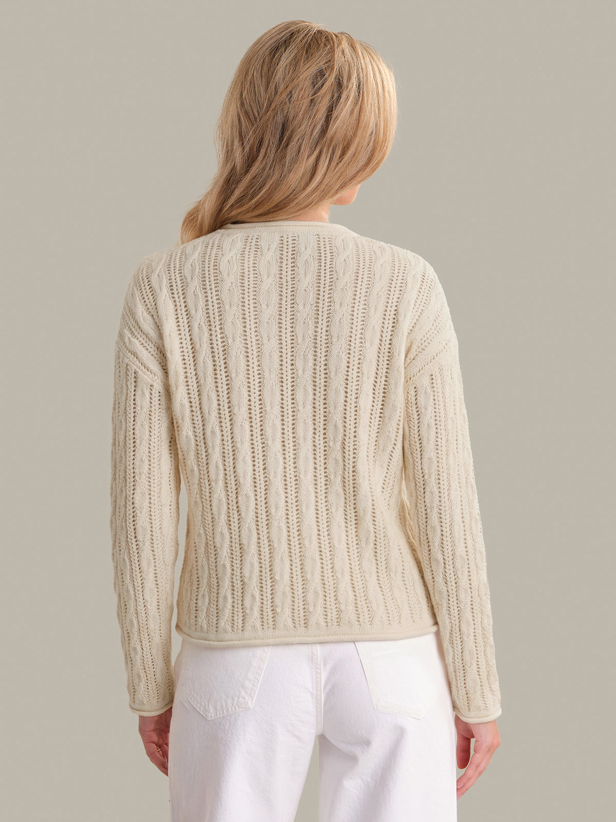 Person wearing a beige knitted sweater and white pants on a plain background