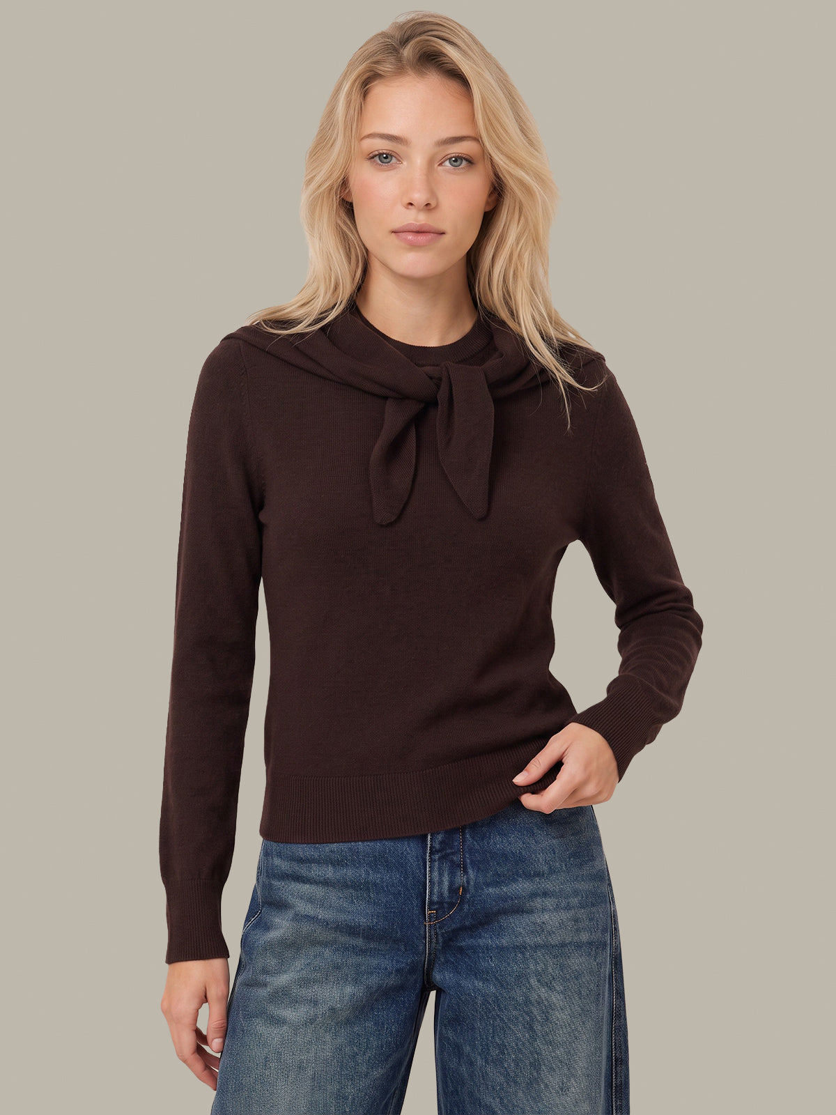 A woman modeling a dark brown long-sleeve knit top with a wide collar and front bow detail, paired with blue jeans, against a solid light gray background.