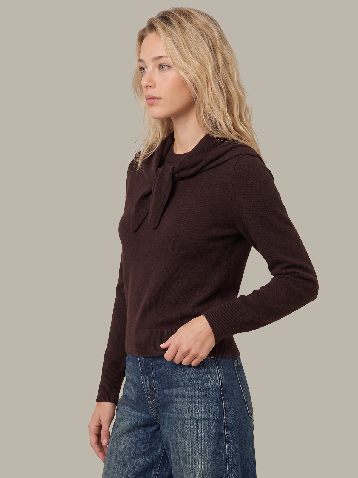 A woman wearing a dark brown long-sleeve knit top with a draped tie-neck detail, paired with blue jeans, shown against a solid neutral gray background.