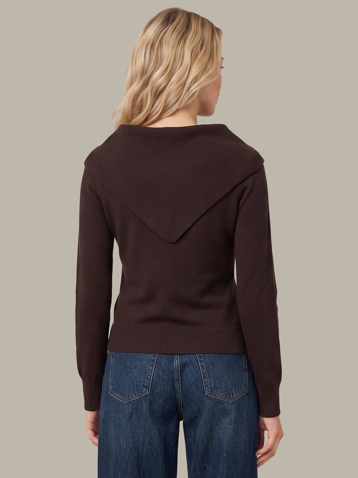 A woman viewed from the back wearing a dark brown knit sweater with a wide draped yoke and blue jeans, set against a solid taupe background.