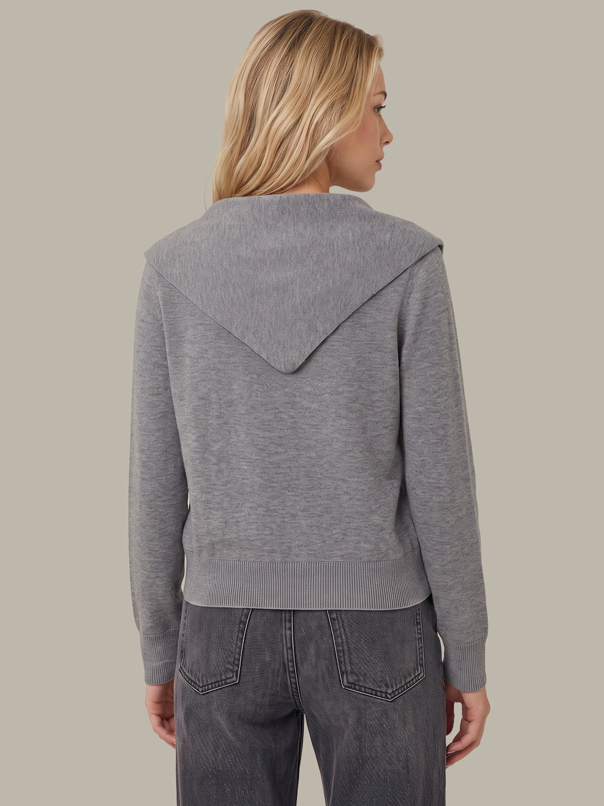 A woman shown from the back wearing a heather gray knit hooded sweater and dark gray jeans, against a solid warm gray studio background.