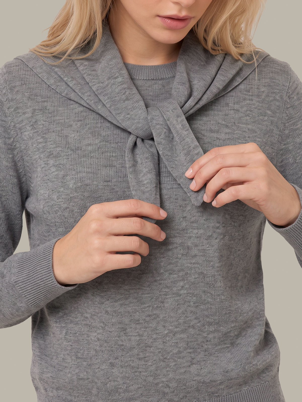 A close-up of a woman wearing a heather gray knit sweater with a wide draped collar tied in a knot at the chest, shown against a plain beige background.