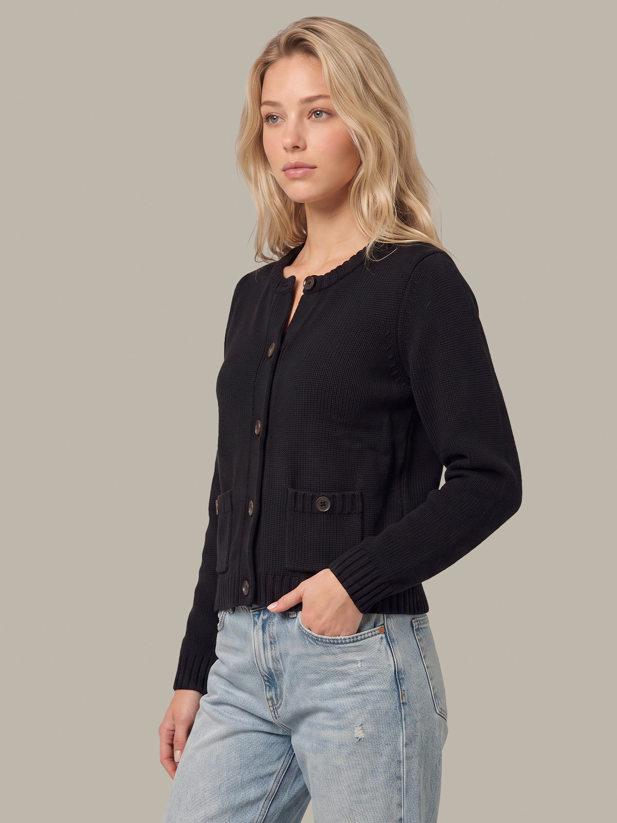 A blonde woman wearing a black long-sleeve button-front cardigan with scalloped trim and light-wash blue jeans, standing against a solid light gray studio background.