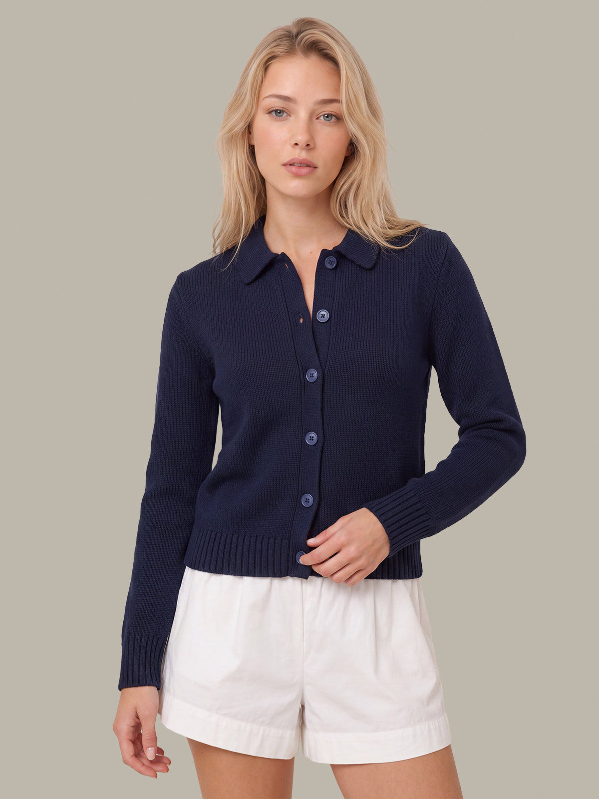 A woman modeling a navy blue ribbed knit cardigan with a button-down front and collar, paired with white shorts, against a solid taupe background.