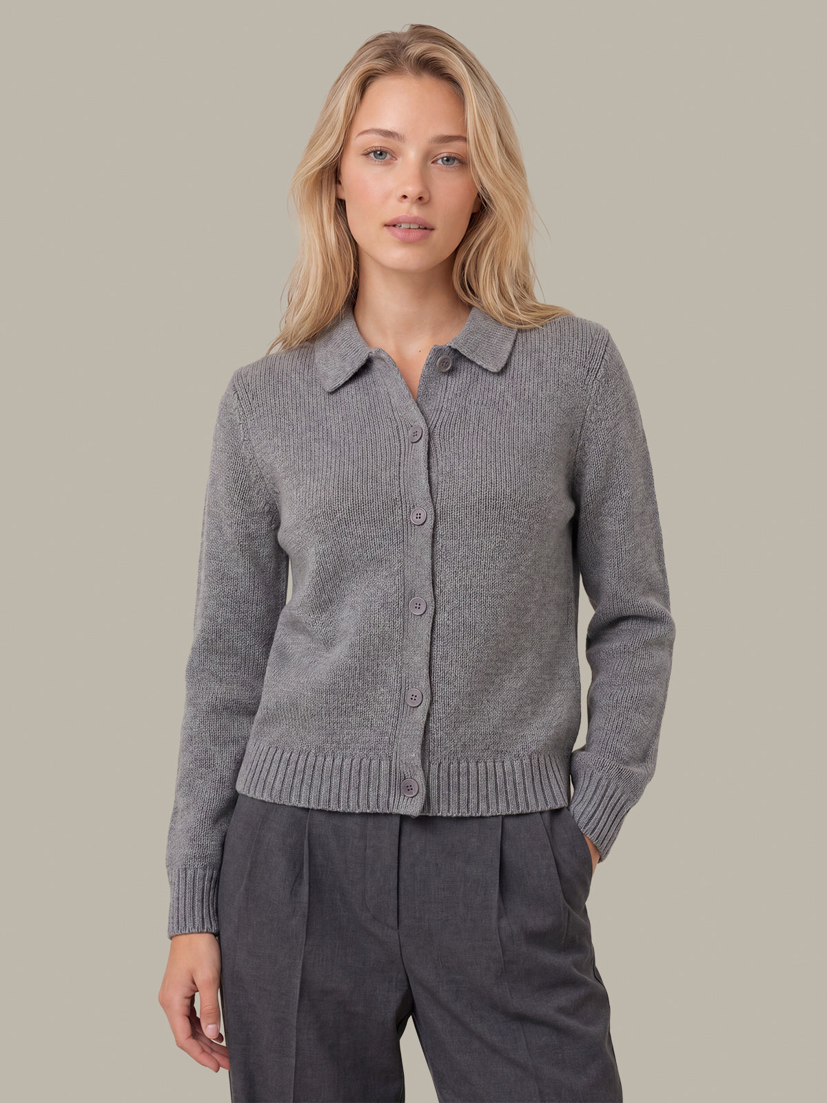 A woman wearing a gray ribbed knit button-front cardigan with a collar, paired with dark gray trousers, against a solid warm gray background.