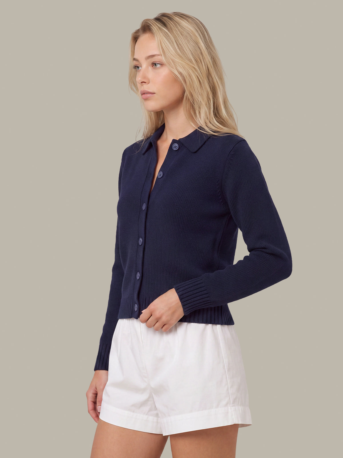 A woman wearing a navy blue knit button-front cardigan with a collar and white shorts, photographed against a plain warm gray studio background.