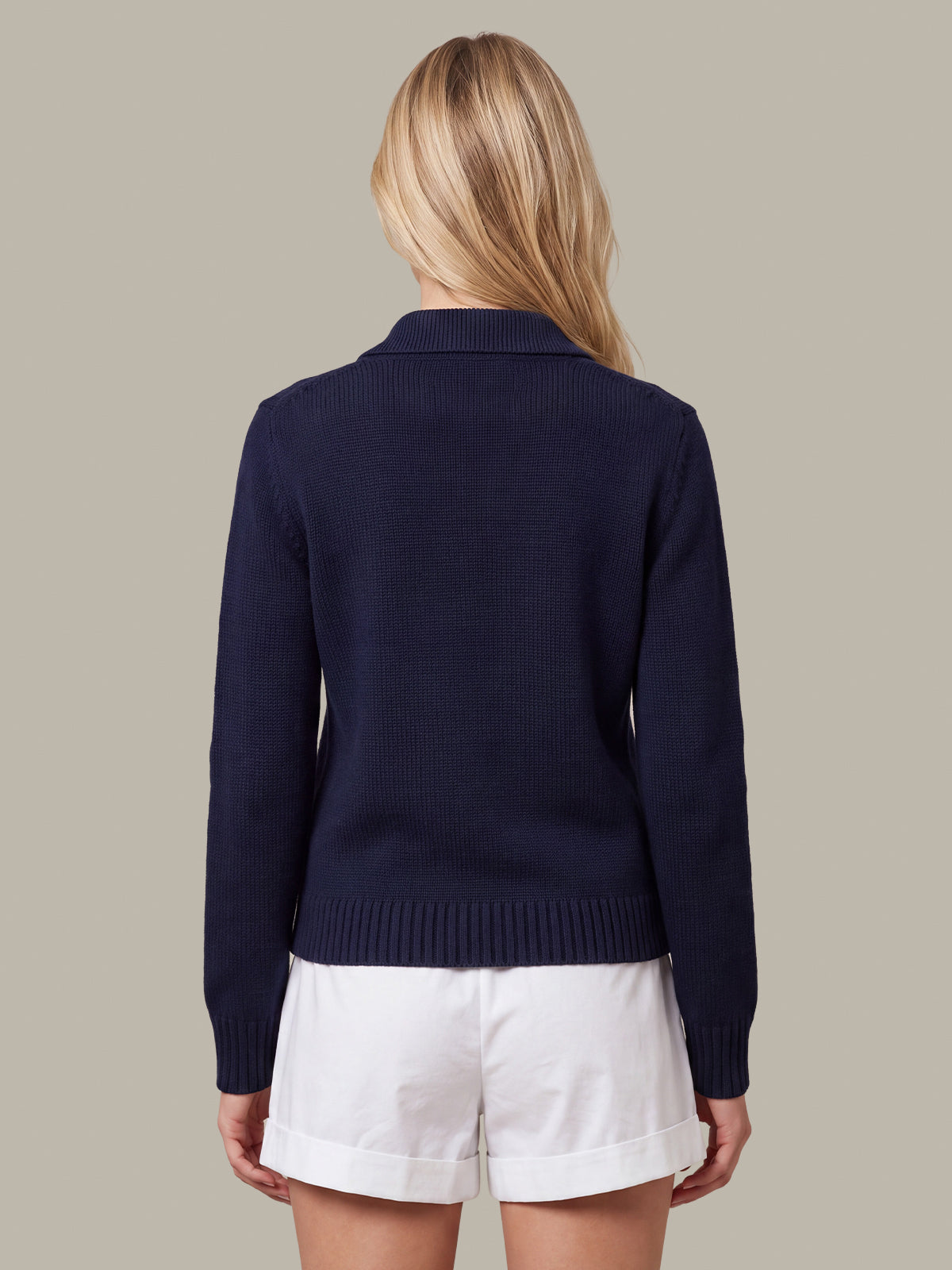 A woman shown from the back wearing a navy blue knit cardigan and white shorts against a solid beige background.