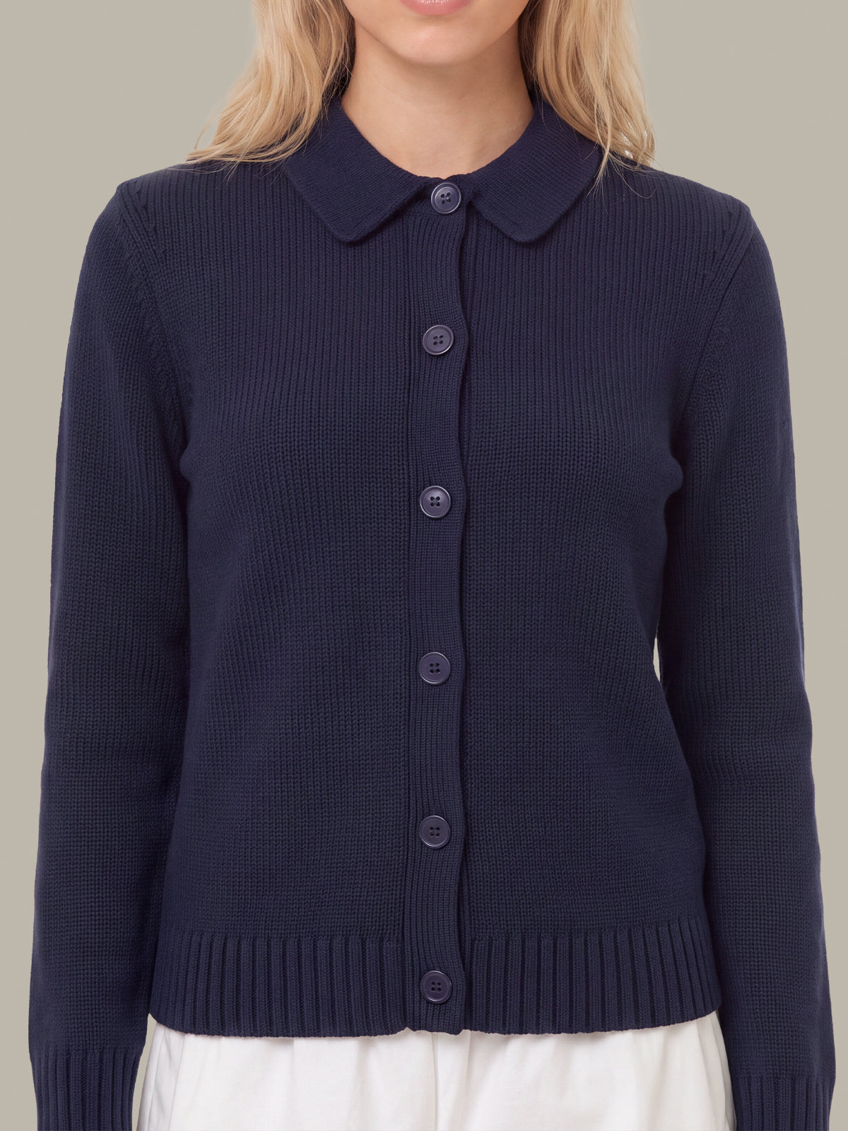 A woman wearing a navy blue ribbed knit cardigan with a Peter Pan collar and front buttons, paired with white bottoms, against a solid beige background.