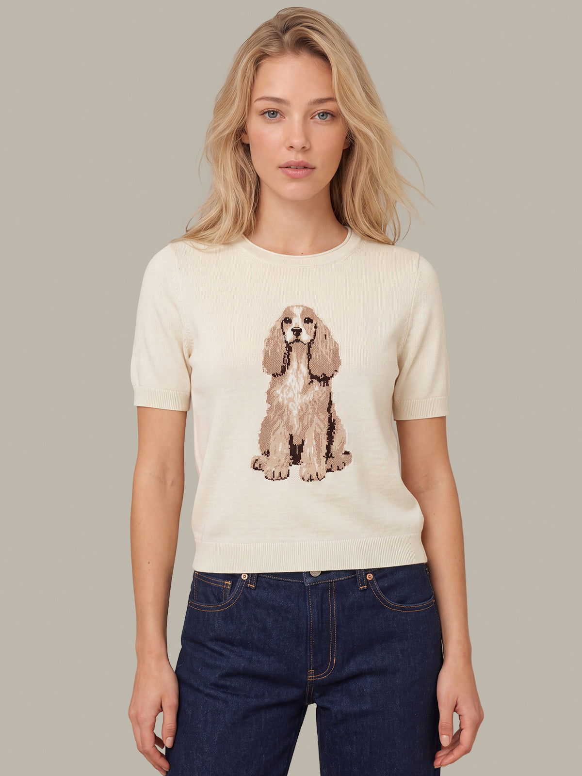 A woman modeling a cream-colored short-sleeve knit sweater featuring a detailed graphic of a Cocker Spaniel dog on the front, paired with dark blue jeans against a solid light gray background.