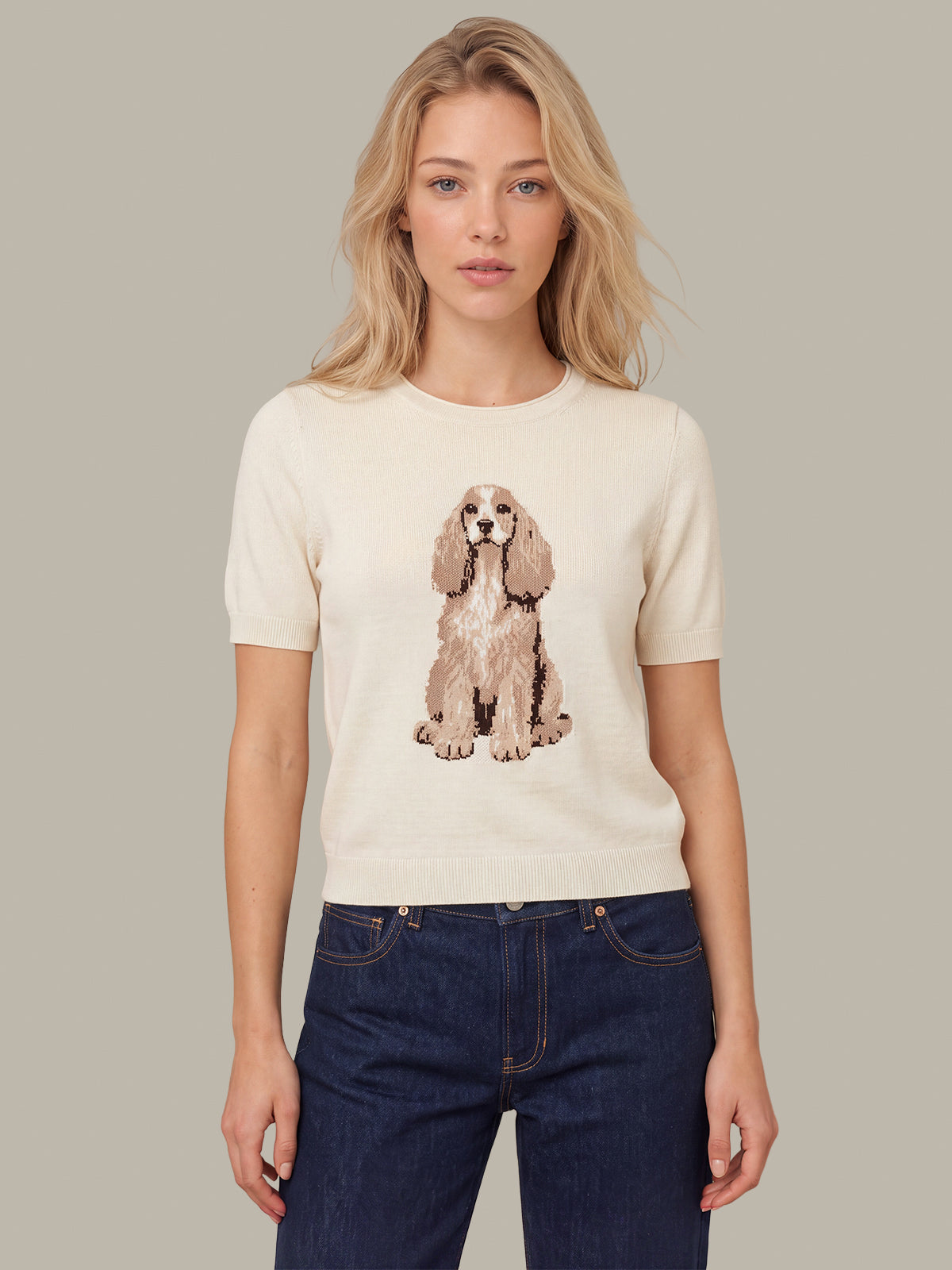 A woman modeling a cream-colored short-sleeve knit sweater featuring a graphic print of a Cocker Spaniel dog on the front, paired with dark blue jeans, against a solid taupe background.