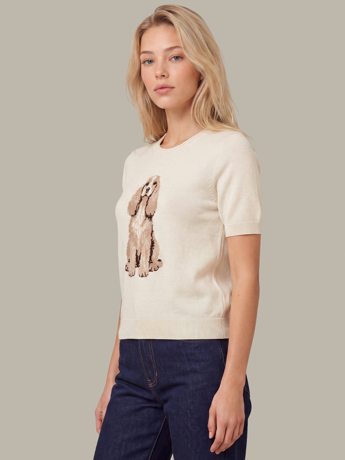 A blonde woman wearing a cream short-sleeve knit sweater with a brown and beige dog graphic on the front, paired with dark blue jeans, against a solid light gray background.