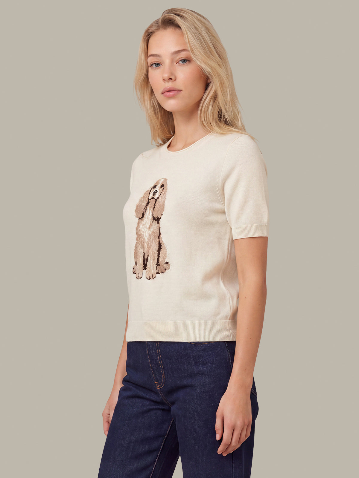 A blonde woman wearing a cream short-sleeve knit sweater with a brown and beige dog graphic on the front, paired with dark blue jeans, set against a solid taupe background.