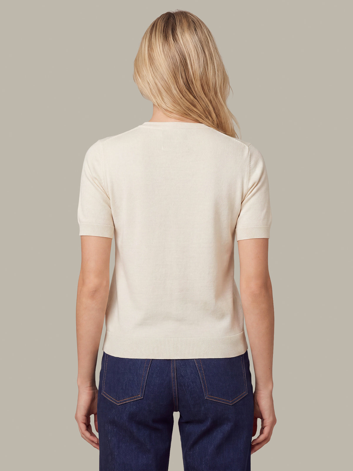 A woman viewed from the back wearing a short-sleeve cream knit top and dark blue jeans, set against a solid taupe background.