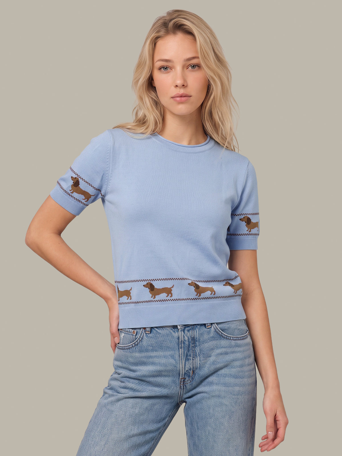 A woman modeling a light blue short-sleeve knit sweater with brown dachshund dog illustrations across the hem and sleeve cuffs, paired with light blue jeans against a solid gray background.